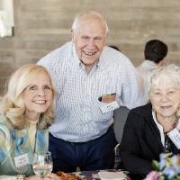 Gentleman standing behind two other event attendees sitting at table, all smiling for camera